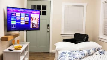 Habitación de hotel con un televisor de pantalla plana montado en la pared que muestra diversos servicios de streaming, una cama con ropa de cama estampada en blanco y azul, una mesita de noche con un tazón de cereal y un vaso de jugo de naranja, y un estante de madera encima de la mesa.