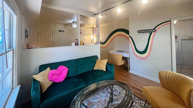 Un salon avec un canapé vert, un fauteuil jaune, une table basse en verre et un mur orné d’un motif ondulé et coloré.