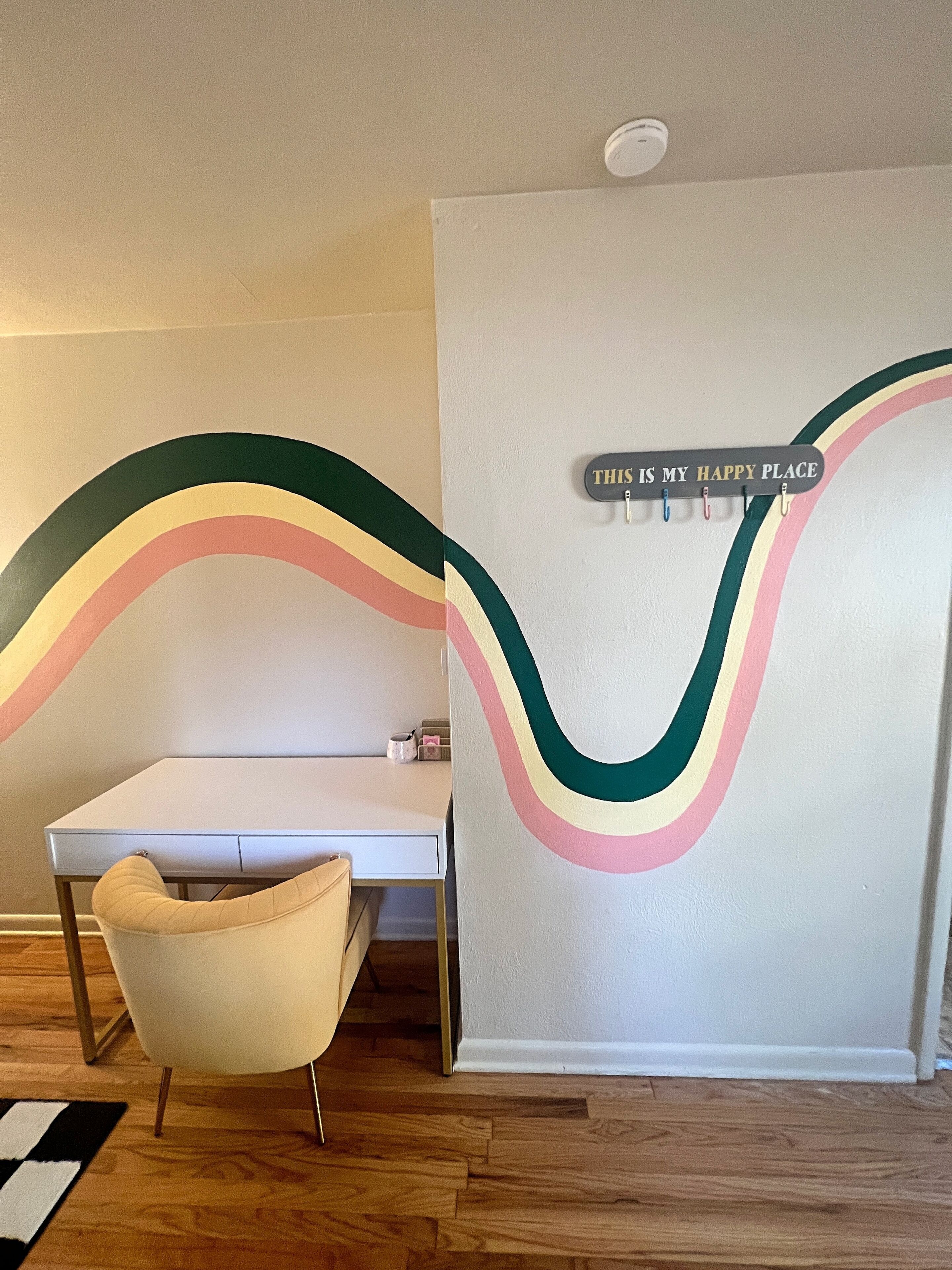 A room with a white desk, a beige chair, and a wall with a rainbow design and a sign that says "THIS IS MY HAPPY PLACE".