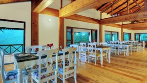 A spacious dining area with white wooden tables and chairs, large windows offering views of a forested area, and a high wooden ceiling. - Dyo - The Organic Village Resort (Dhari)