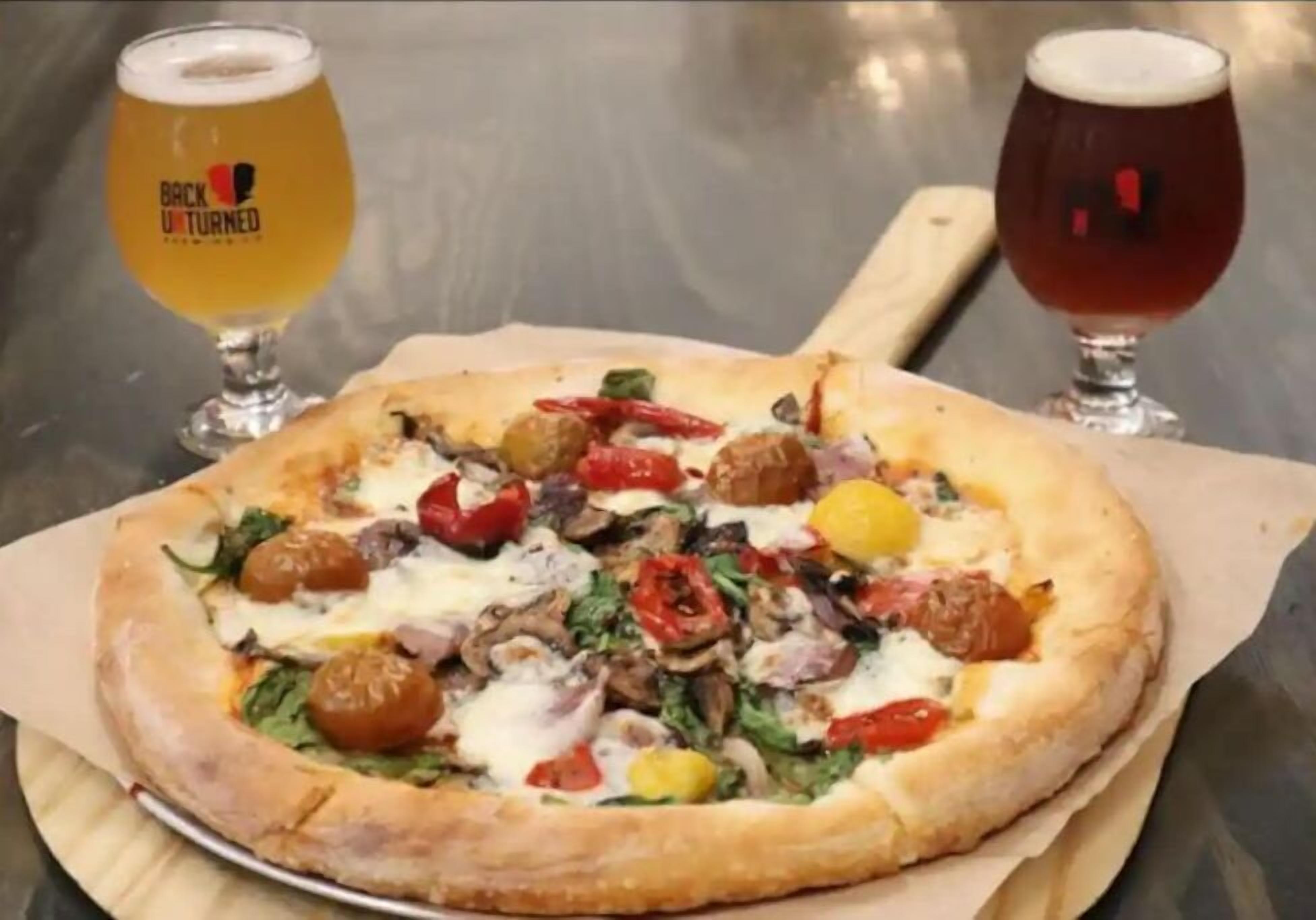 Pizza met diverse toppings op een houten plank, vergezeld van twee glaasjes bier.