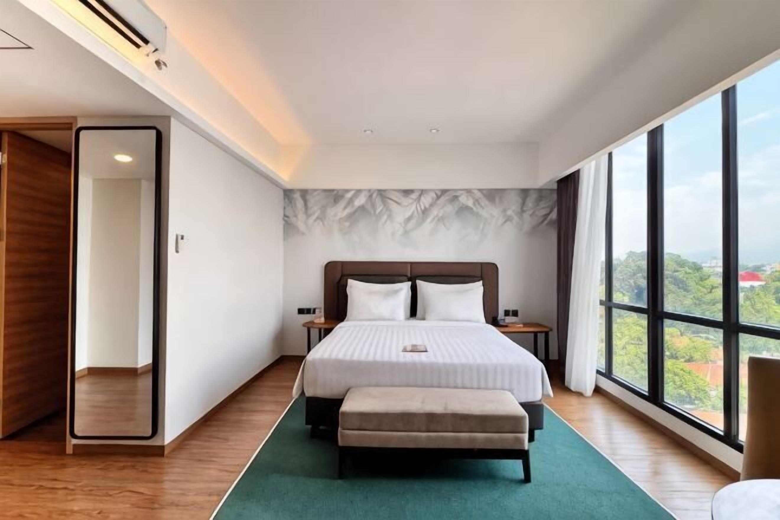 A modern hotel room with a large bed, a green rug, a wooden floor, a nightstand, and a view of the city through large windows.