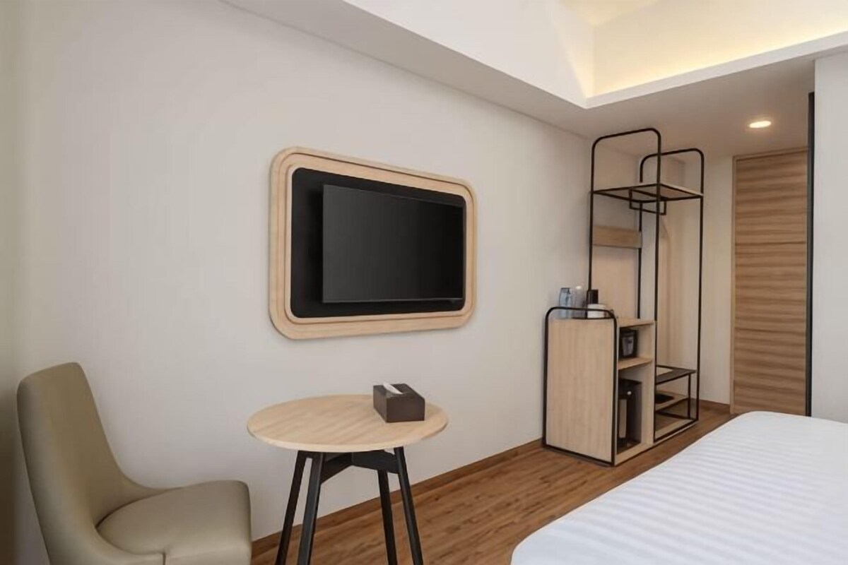 A modern hotel room with a bed, a desk, a chair, a television, and a wardrobe.