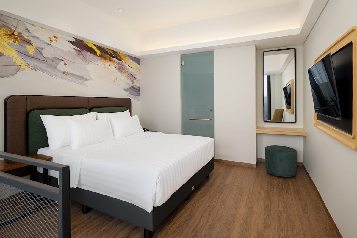 A modern hotel room with a large bed, a flat-screen TV, a mirror, and a green ottoman.