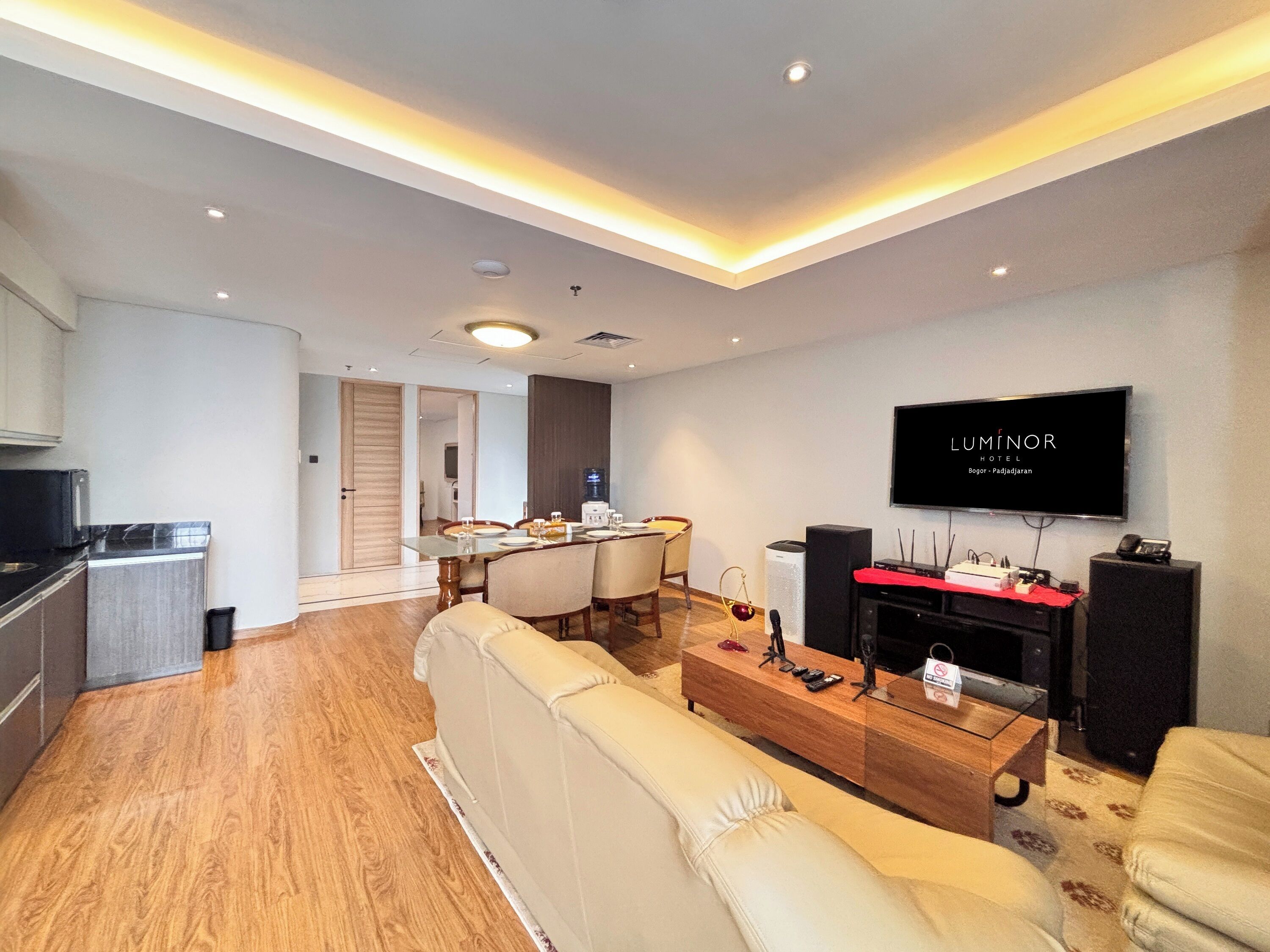 A modern hotel living room with a sofa, a coffee table, a TV, and a dining area with a table and chairs.