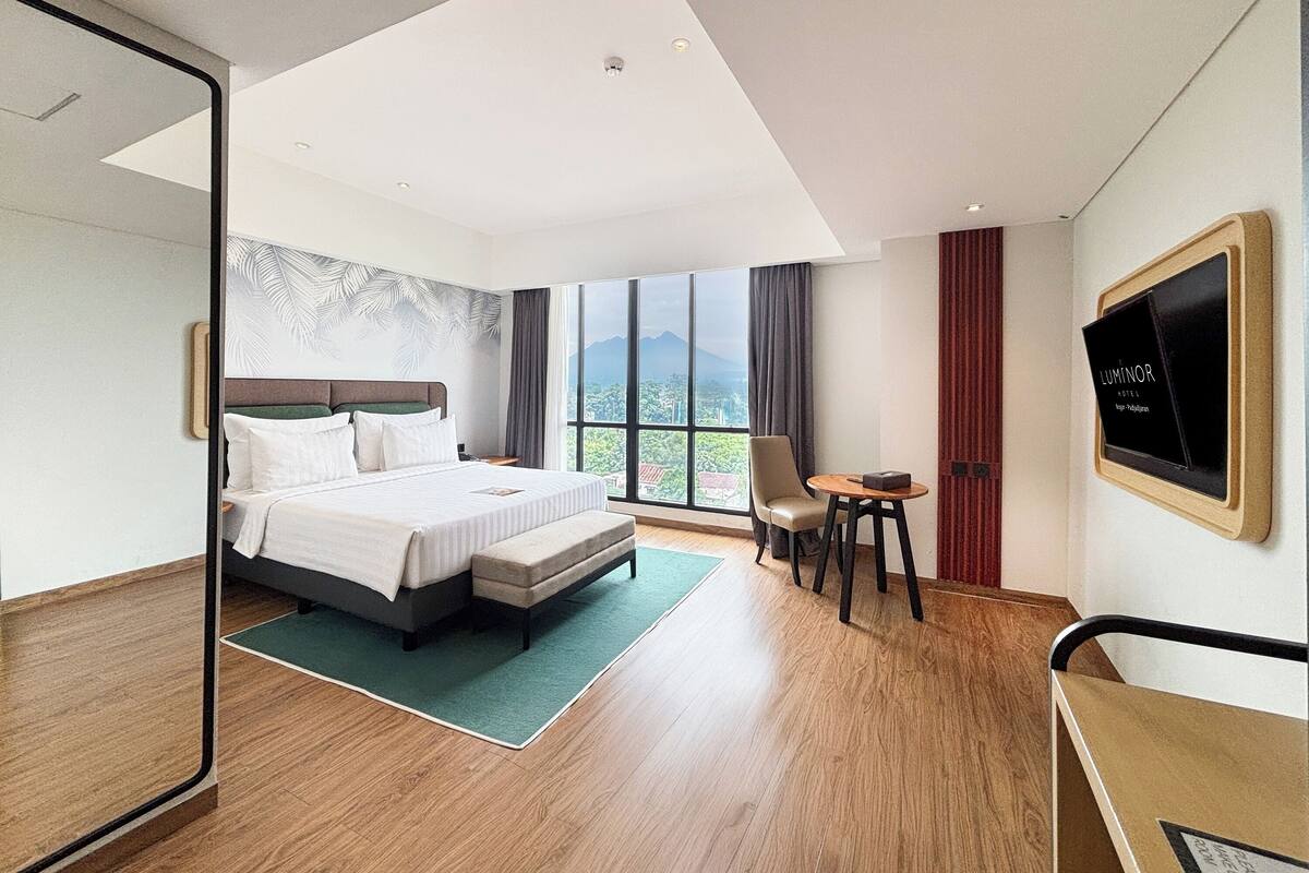 A modern hotel room with a large bed, a seating area, a TV, and a view of the city and mountains.