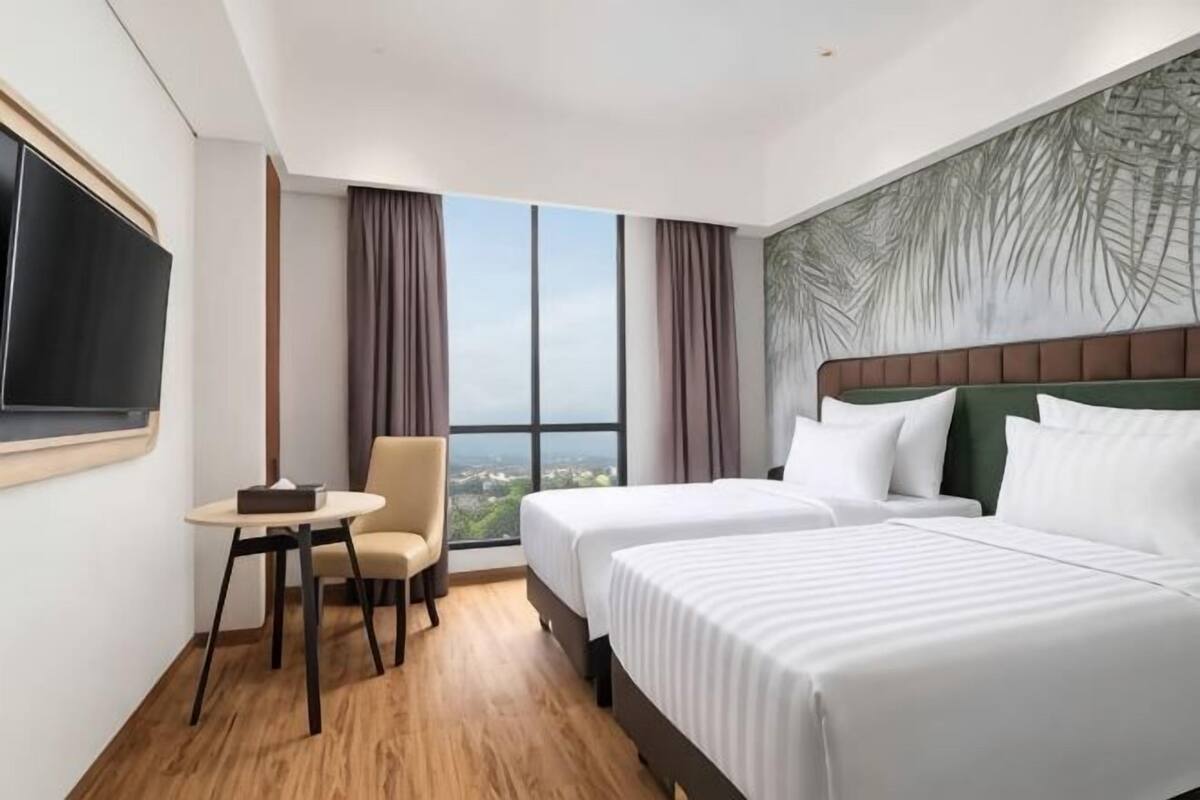 A hotel room with two beds, a desk, a chair, a large window with curtains, and a flat-screen TV.