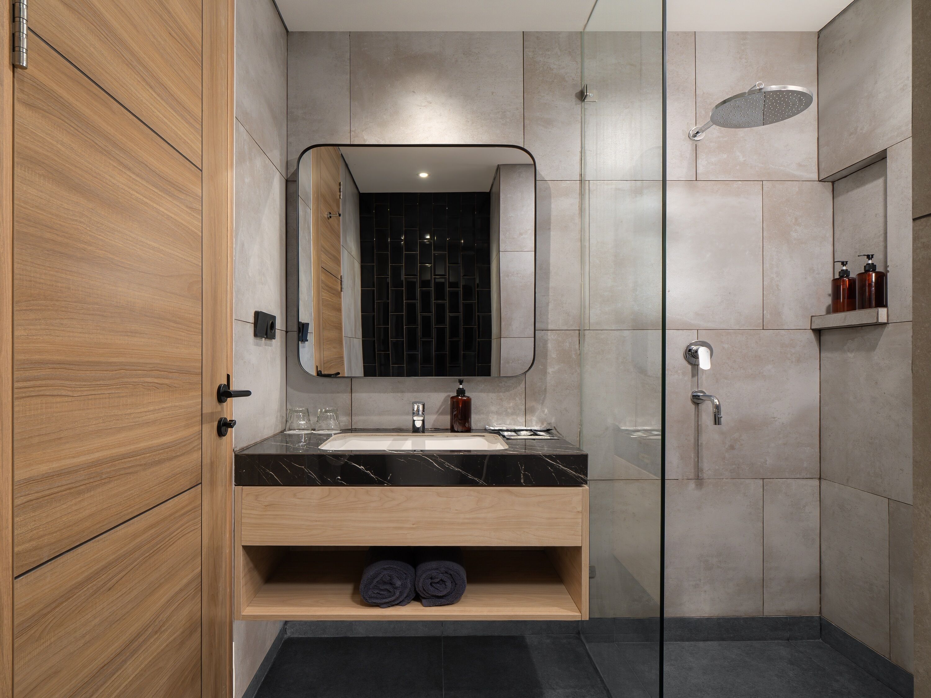 Une salle de bain moderne avec un meuble-lavabo en bois, un grand miroir, une cabine de douche en verre et une zone de douche carrelée de couleur sombre.