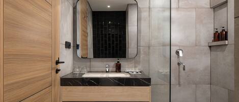 Un baño moderno con tocador de madera, un espejo grande, cabina de ducha de vidrio y zona de ducha con azulejos oscuros.