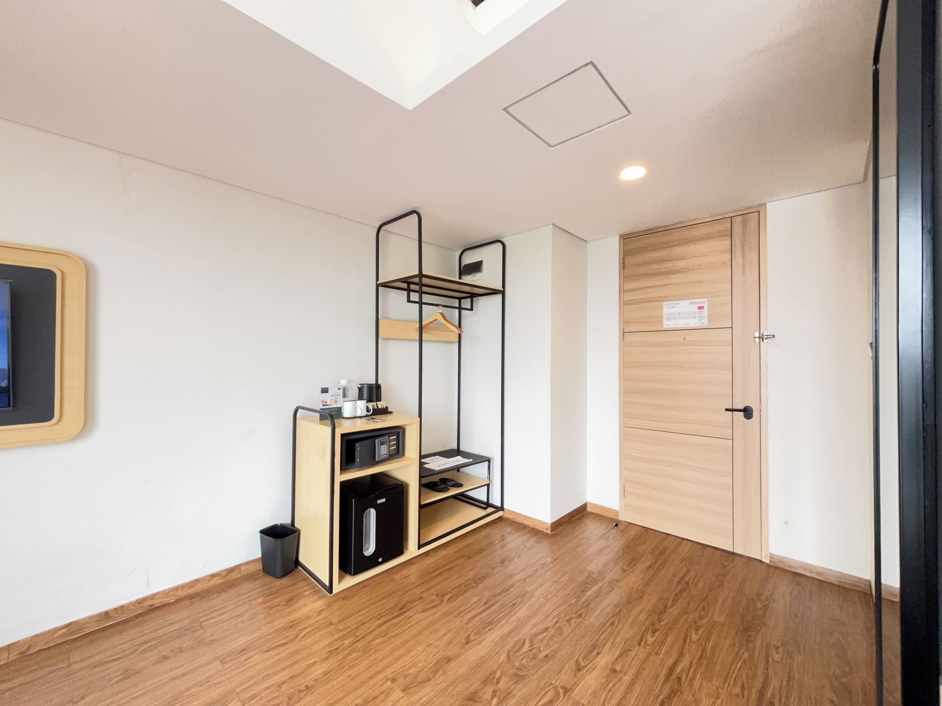 A modern hotel room with a wooden floor, a black-framed shelf unit with a coffee maker, a microwave, and a small trash bin.