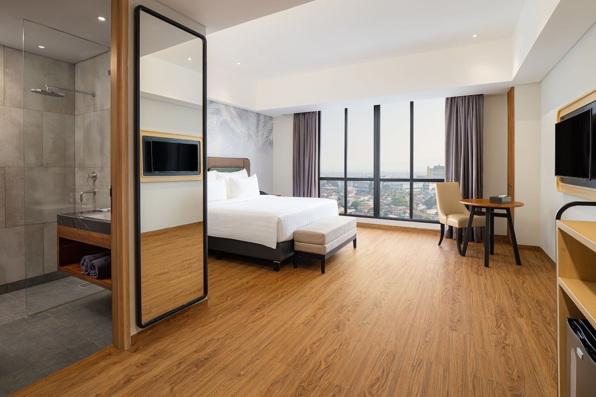 A modern hotel room with a large bed, a sitting area with a TV, a bathroom with a shower, and a city view.