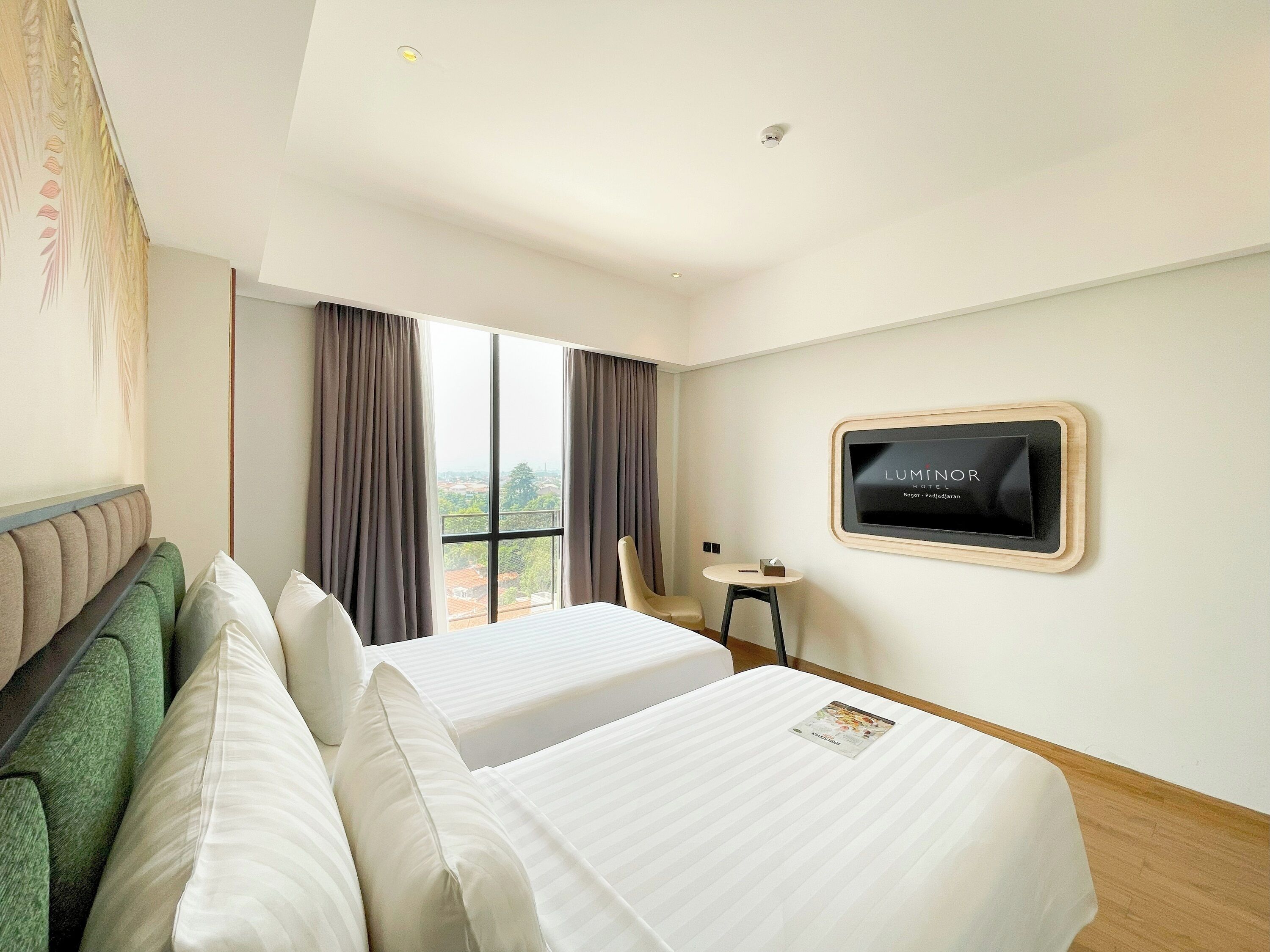 A hotel room with two beds, a small table, a chair, a TV on the wall, and a view of the city through the window.