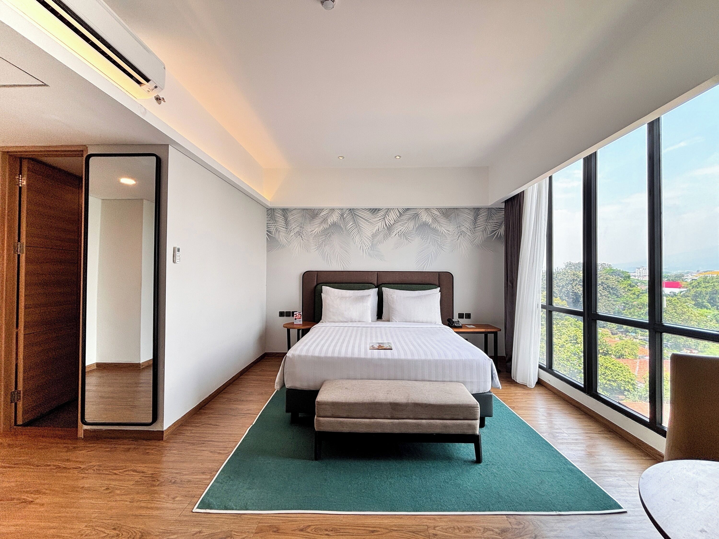 A modern hotel room with a large bed, a bench, a view of the city, and a wooden floor.
