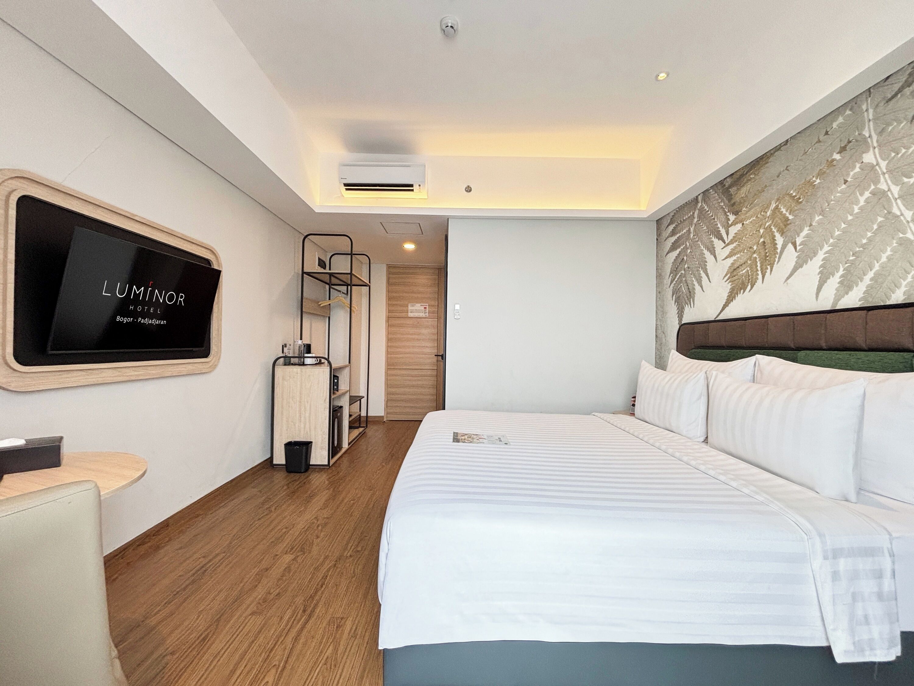A hotel room with a bed, a TV mounted on the wall, a wooden floor, and a wall-mounted air conditioner.