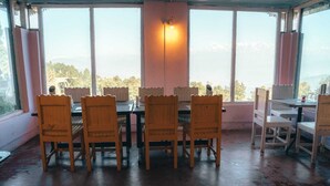 Un comedor con sillas y mesas de madera, ventanas amplias con vista a un paisaje montañoso, y una lámpara fijada a la pared.