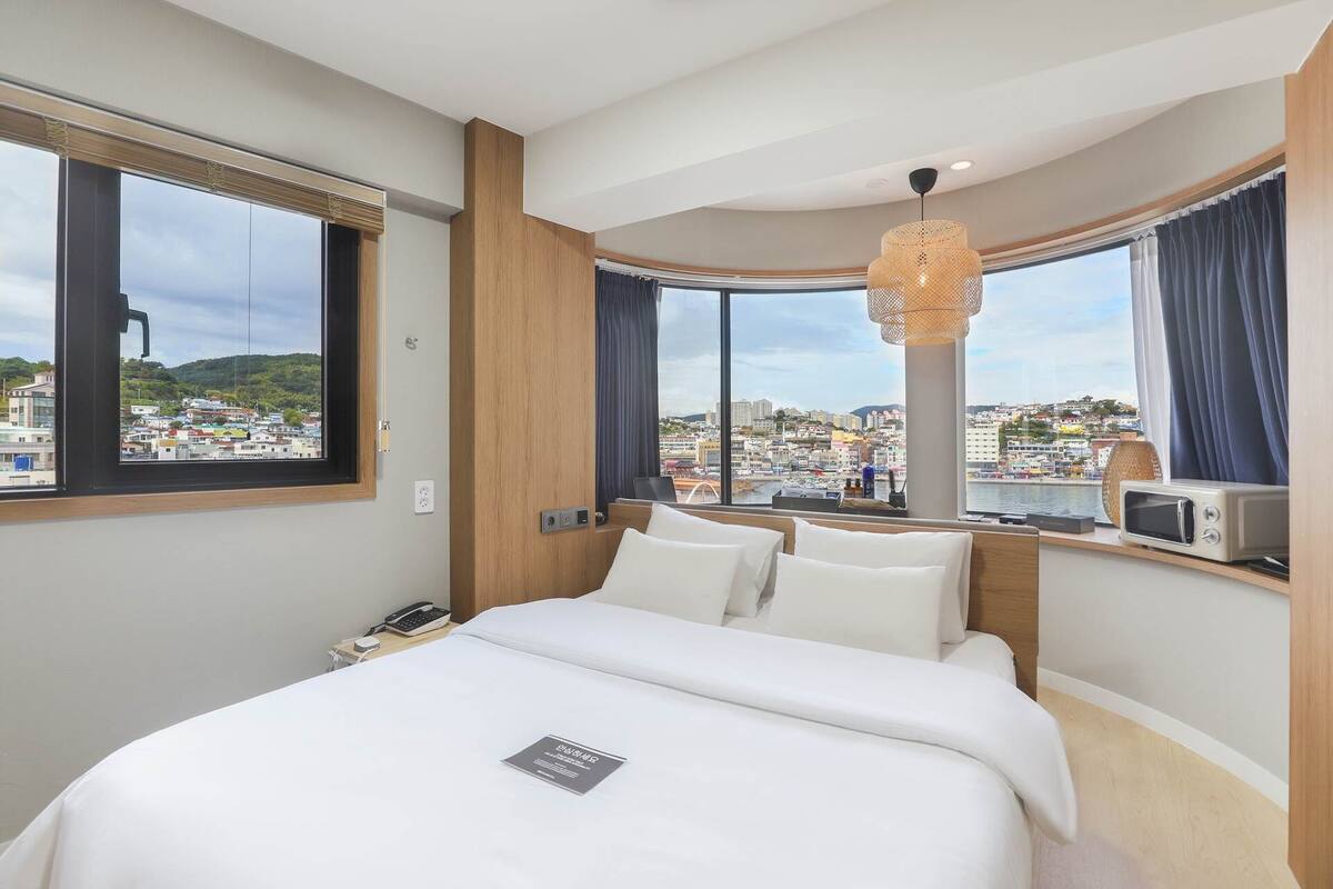 A hotel room with a large bed, a desk with a computer, a telephone, and a view of a cityscape.