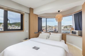 A hotel room with a large bed, a desk with a computer, a telephone, and a view of a cityscape. - DELAMOUR Hotel (Tongyeong)