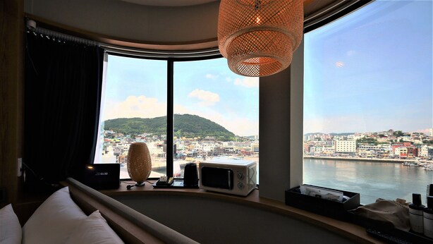 A hotel room with a large window, a bed, a lamp, a microwave, and a small table with items on it. - DELAMOUR Hotel (Tongyeong)