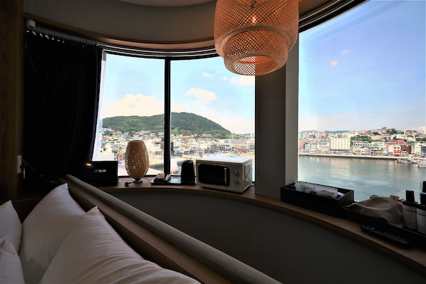A hotel room with a large window, a bed, a lamp, a microwave, and a small table with items on it. - DELAMOUR Hotel (Tongyeong)