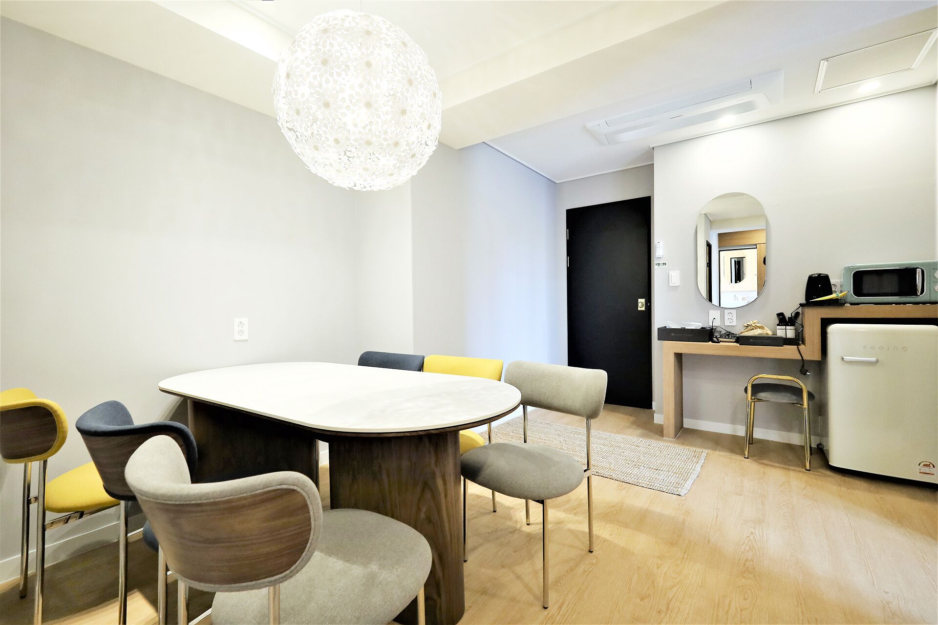 A modern hotel room with a round dining table, multiple chairs, a small kitchenette, a microwave, and a ceiling light fixture.