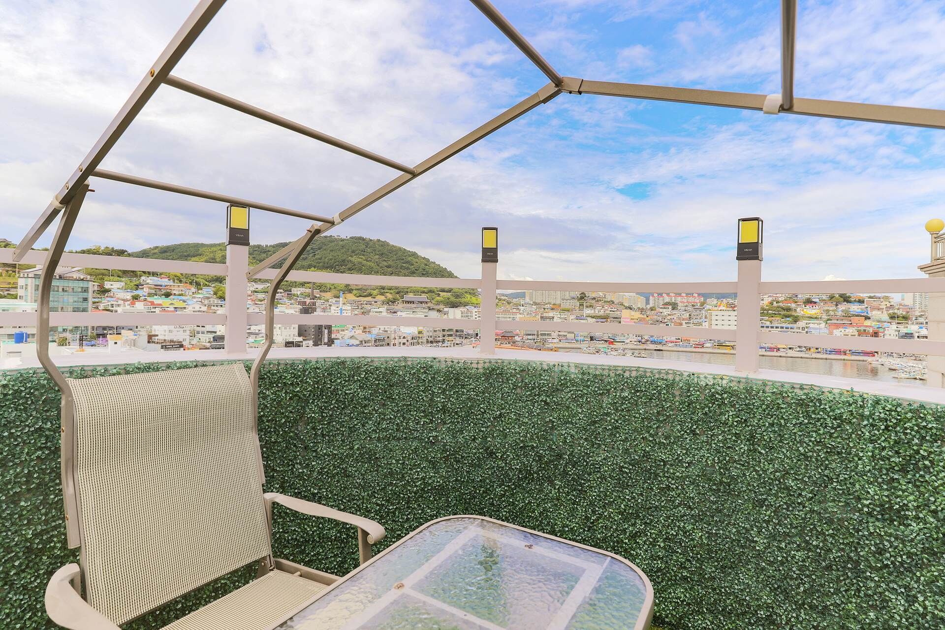 Una silla con respaldo tejido y una mesa de vidrio, frente a un paisaje urbano y una colina verde.