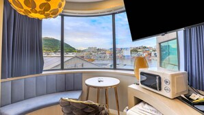 A room with a curved bench, a small round table, a microwave, and a large window offering a city view. - DELAMOUR Hotel (Tongyeong)