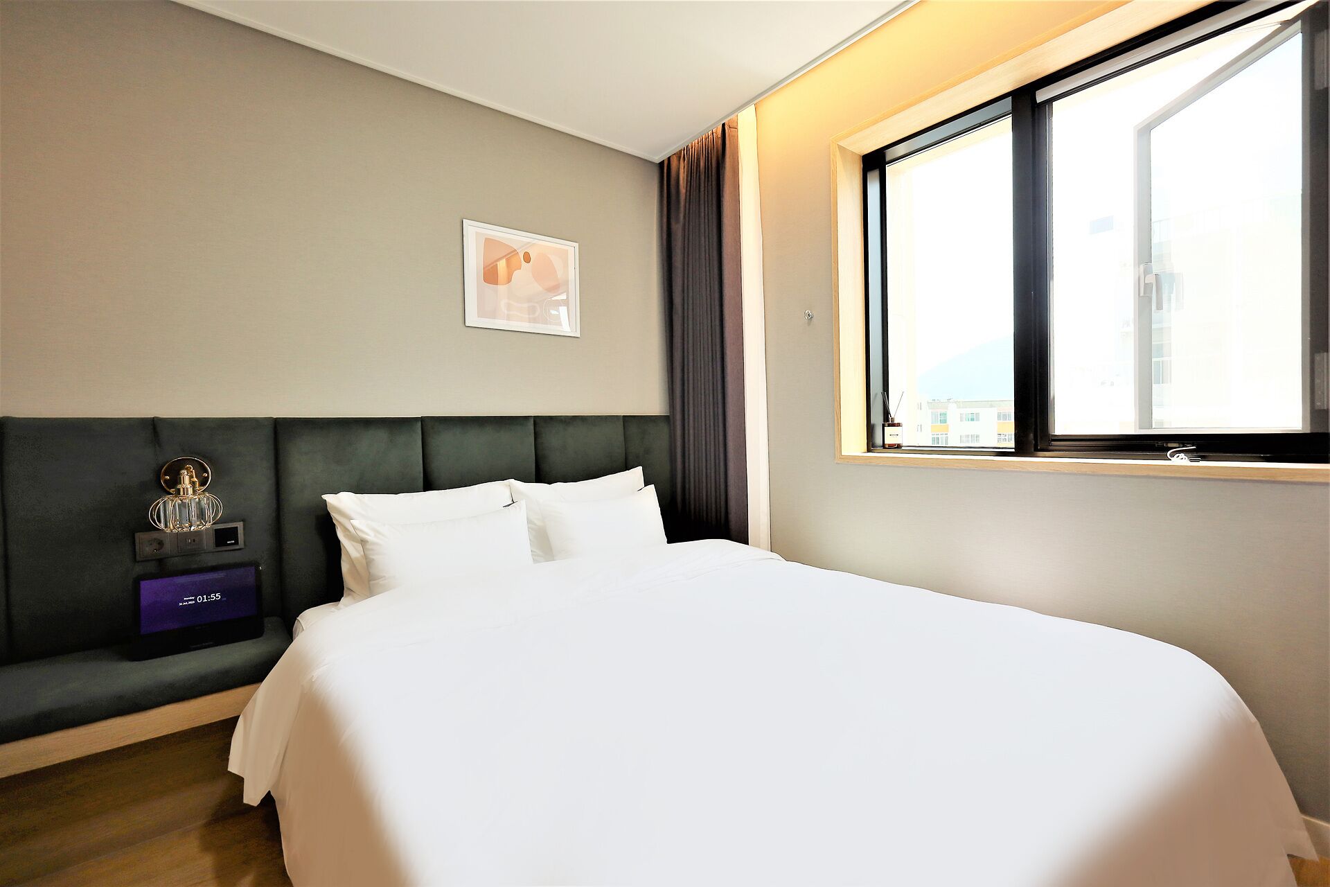 A hotel room with a bed, a bedside table with a lamp, a window with curtains, and a wall-mounted artwork.