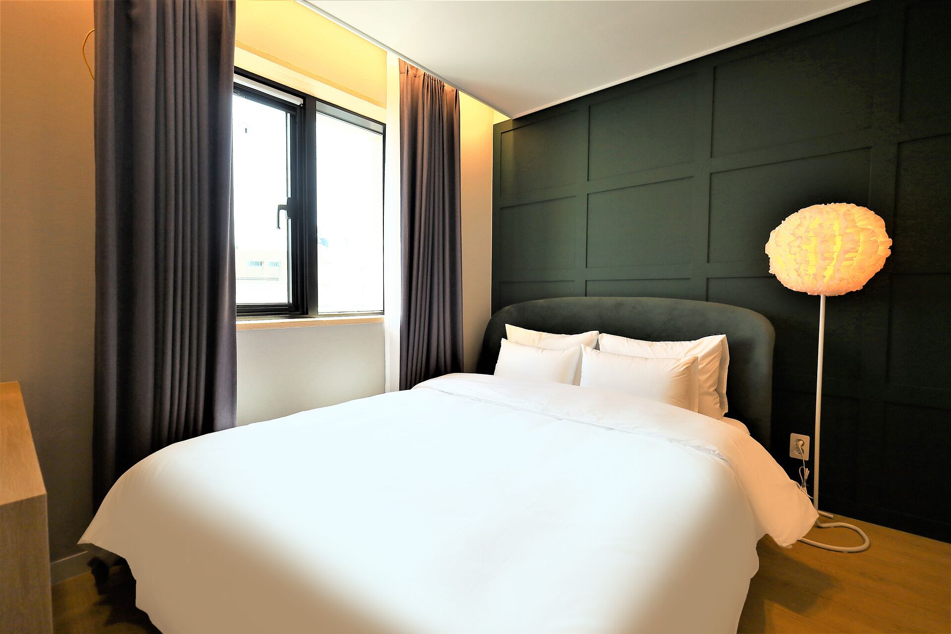 Hotelkamer met een bed, een raam met gordijnen, een wandlamp en een donkerbruin hoofdeinde.