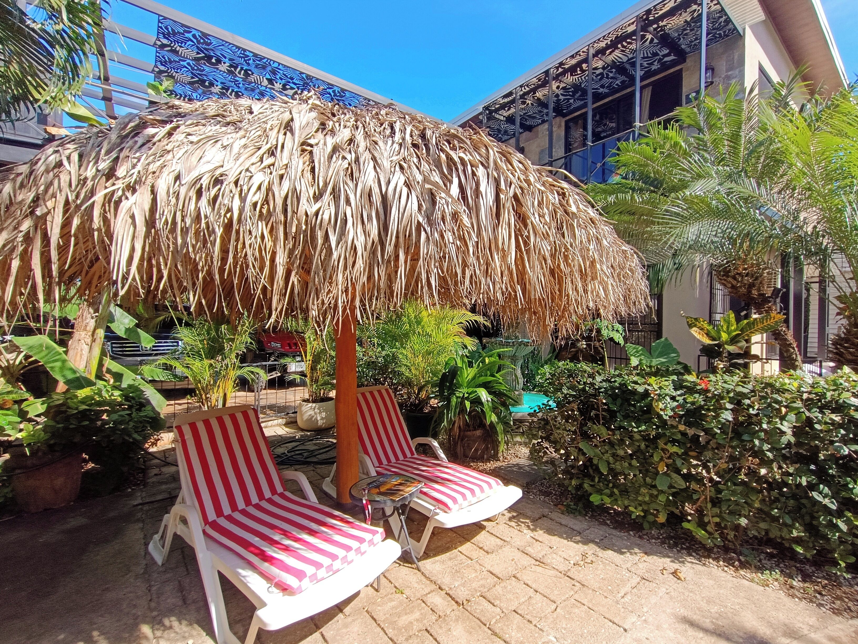 A patio area with two striped lounge chairs under a thatched umbrella, surrounded by potted plants and a pool.