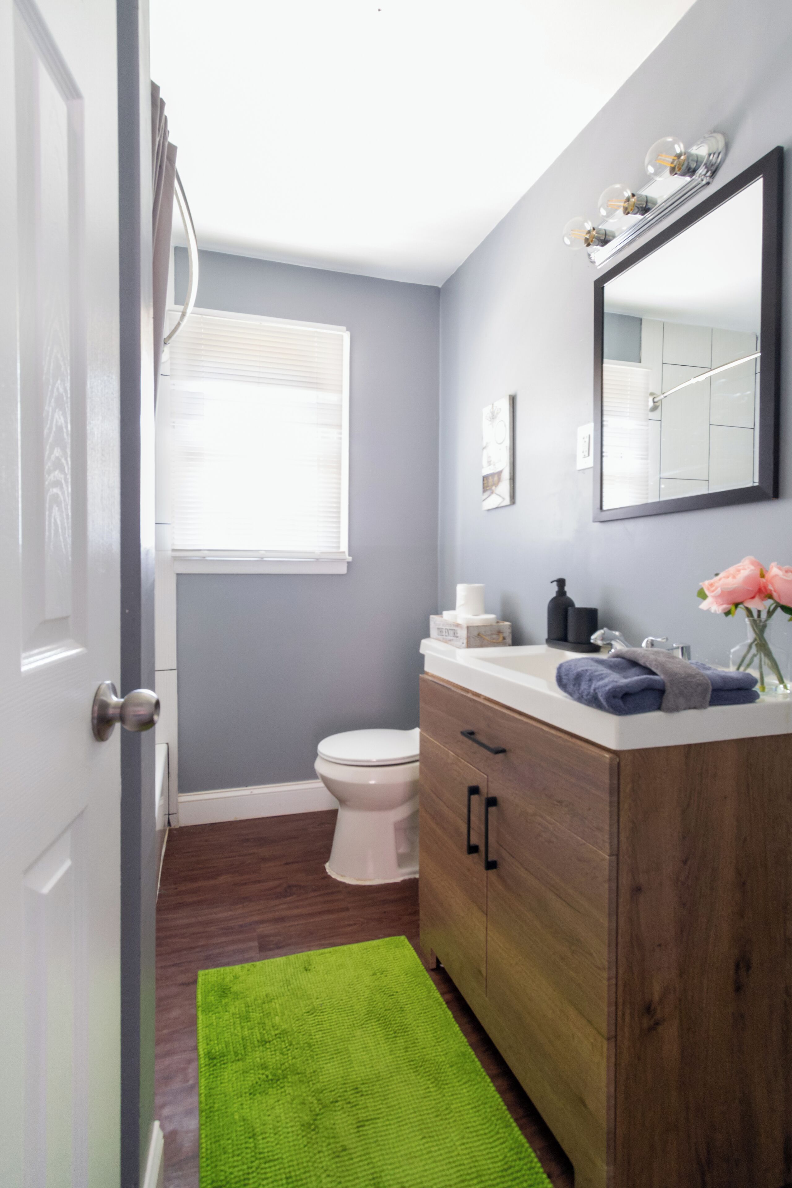 Un baño con tocador de madera, inodoro blanco, alfombra verde y ventana con persianas.