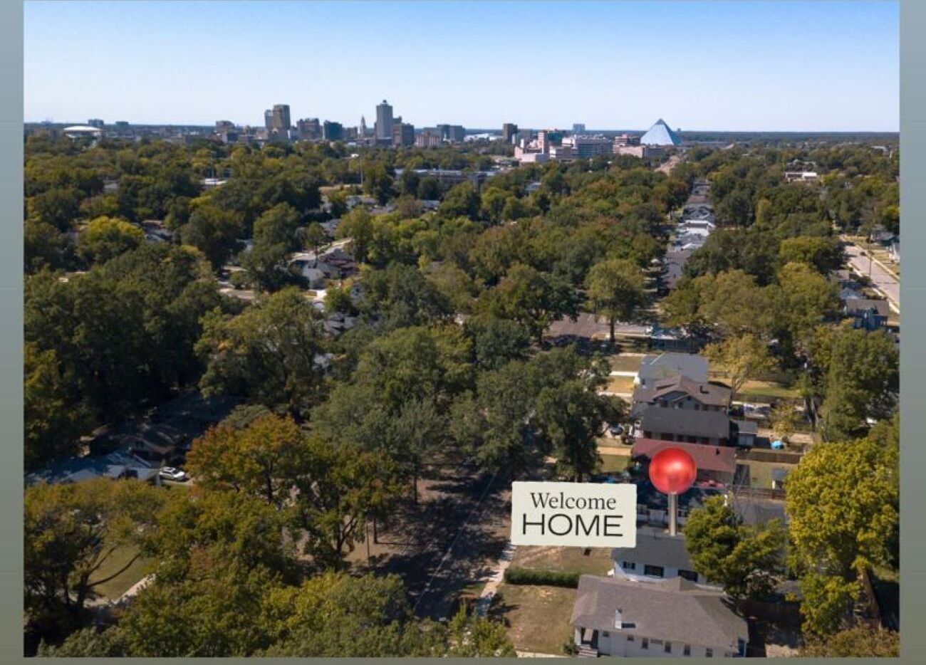 An aerial view of a residential area with a prominent 'Welcome HOME' sign, surrounded by lush greenery and a mix of single and multi-story houses.