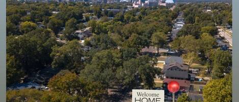 An aerial view of a residential area with a prominent 'Welcome HOME' sign, surrounded by lush greenery and a mix of single and multi-story houses.