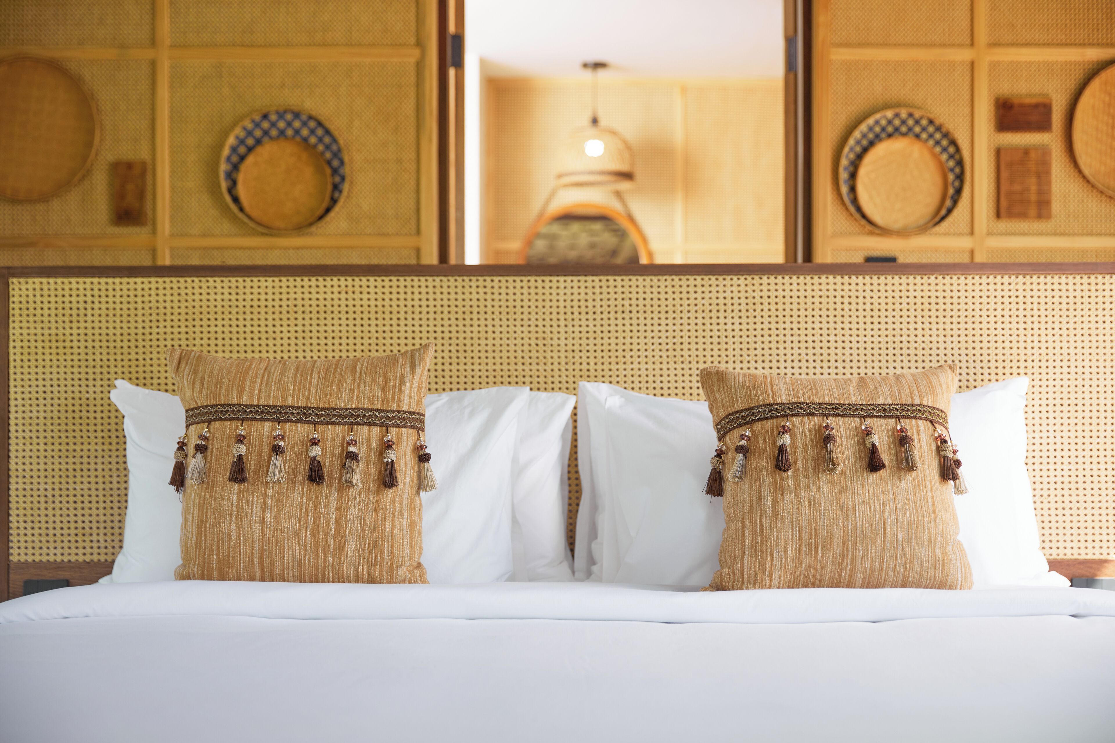 A hotel room with a bed featuring two decorative pillows with tassels, a woven headboard, and a large mirror reflecting the room's decor.