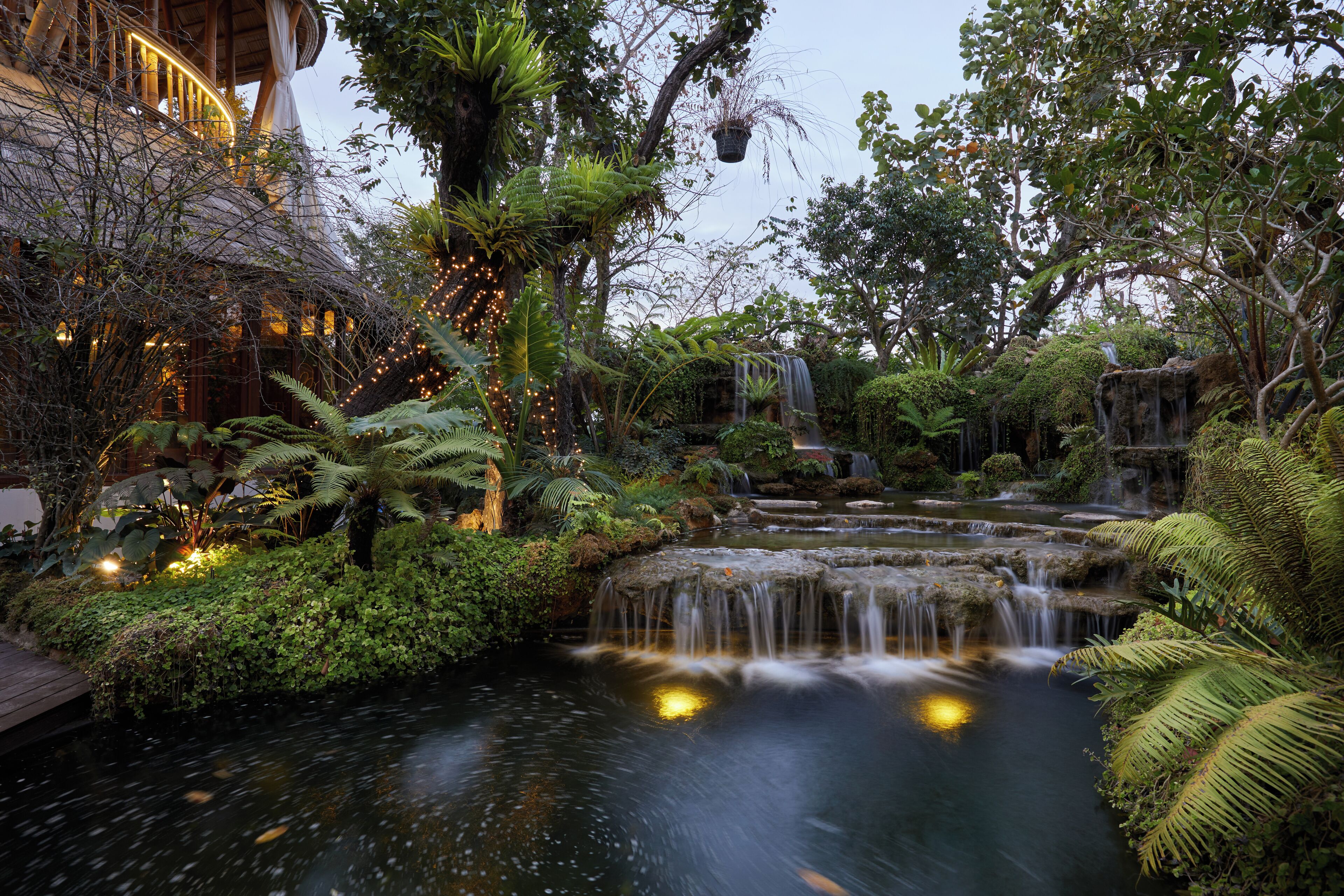 A lush garden with a waterfall, a wooden deck, and string lights.