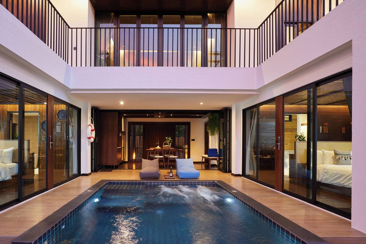 Un área de piscina interior moderna con una barrera de vidrio, suelo de madera y una zona de estar con sillas y una mesa.