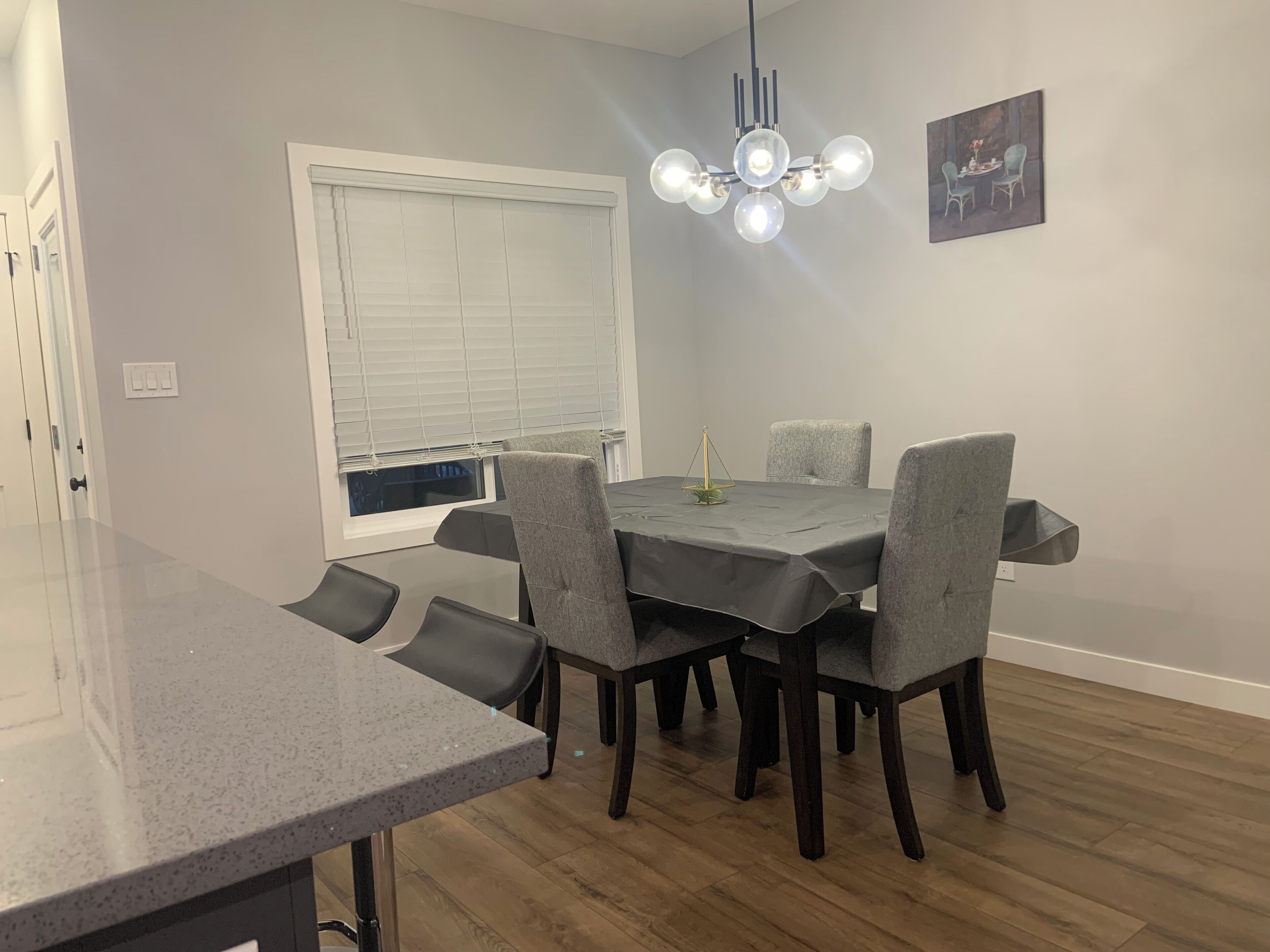 Un coin repas comprenant une table, des chaises, un lustre et une fenêtre avec des stores.