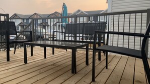 A wooden deck with a glass-top table and black metal chairs, overlooking a row of houses with white siding and gabled roofs.