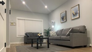 A modern living room with a grey sofa, two armchairs, a coffee table, and a large window with blinds. There are two framed artworks on the wall and a floor lamp.