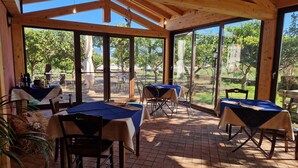 A covered outdoor dining area with tables and chairs, large windows, and a view of trees and greenery. - Lumia (Riposto)
