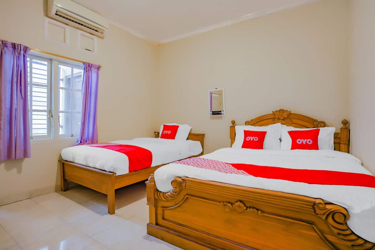 A hotel room with two wooden beds, each with white bedding and red accents, a window with purple curtains, and a wall-mounted air conditioning unit.