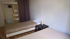 Ein schlichtes Zimmer mit zwei Einzelbetten, einem kleinen Tisch mit Lampe, einem Ventilator und einer Holzleiter mit zusammengeklappten Handtüchern.