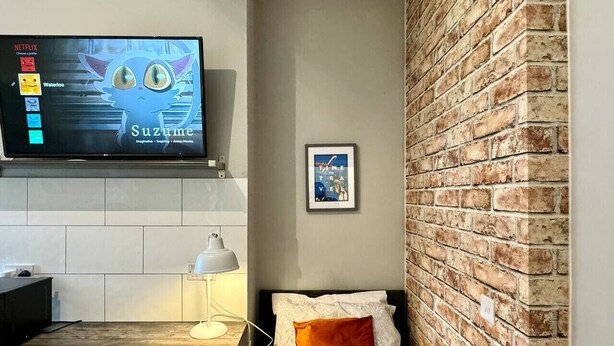 Una habitación pequeña con cama, escritorio, televisor mostrando una caricatura y pared de ladrillo.