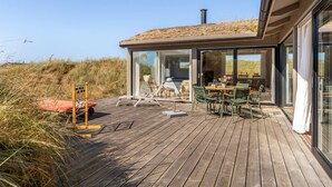 A modern outdoor deck with a wooden floor, glass doors leading to a seating area with chairs and a table, and a view of the surrounding landscape. - "Hoat" - 300m from the sea by Interhome (Saltum)
