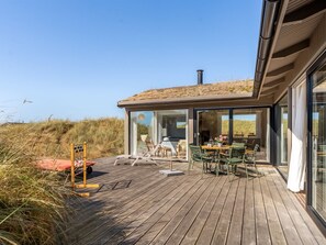 A modern outdoor deck with a wooden floor, glass doors leading to a seating area with chairs and a table, and a view of the surrounding landscape.