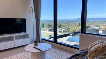 A modern living room with a large TV, a round side table, and a view of a cityscape through large windows.
