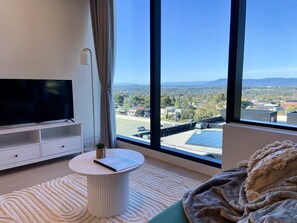 A modern living room with a large TV, a round side table, and a view of a cityscape through large windows.
