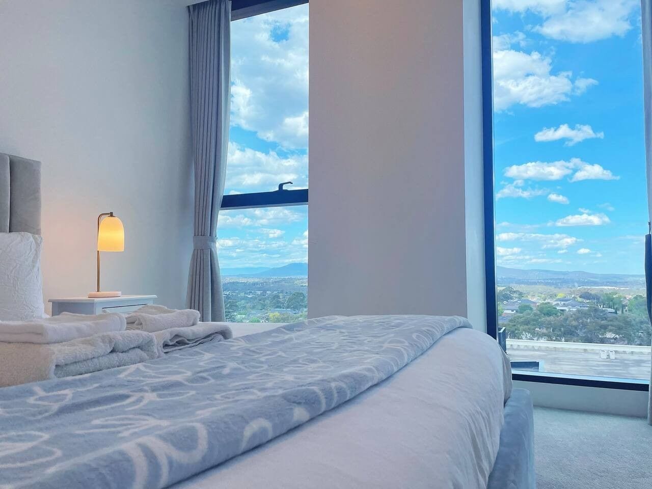 A hotel room with a large bed, a nightstand, a lamp, and a view of the city through a large window.