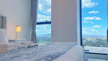 A hotel room with a large bed, a nightstand, a lamp, and a view of the city through a large window.