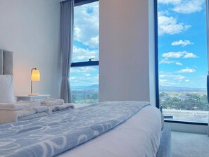 A hotel room with a large bed, a nightstand, a lamp, and a view of the city through a large window.