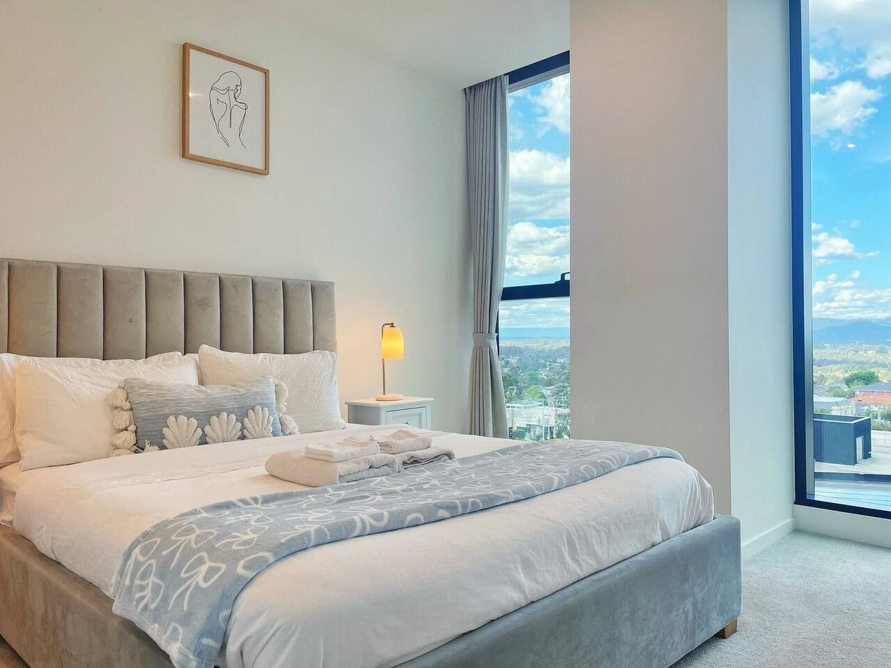 A hotel room with a large bed, a nightstand, a lamp, and a view of the city.